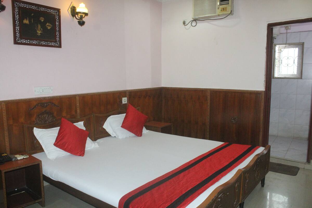 A hotel room with a wooden headboard, a bed with white bedding and red pillows, a bedside table with a lamp, and a wall-mounted air conditioning unit.