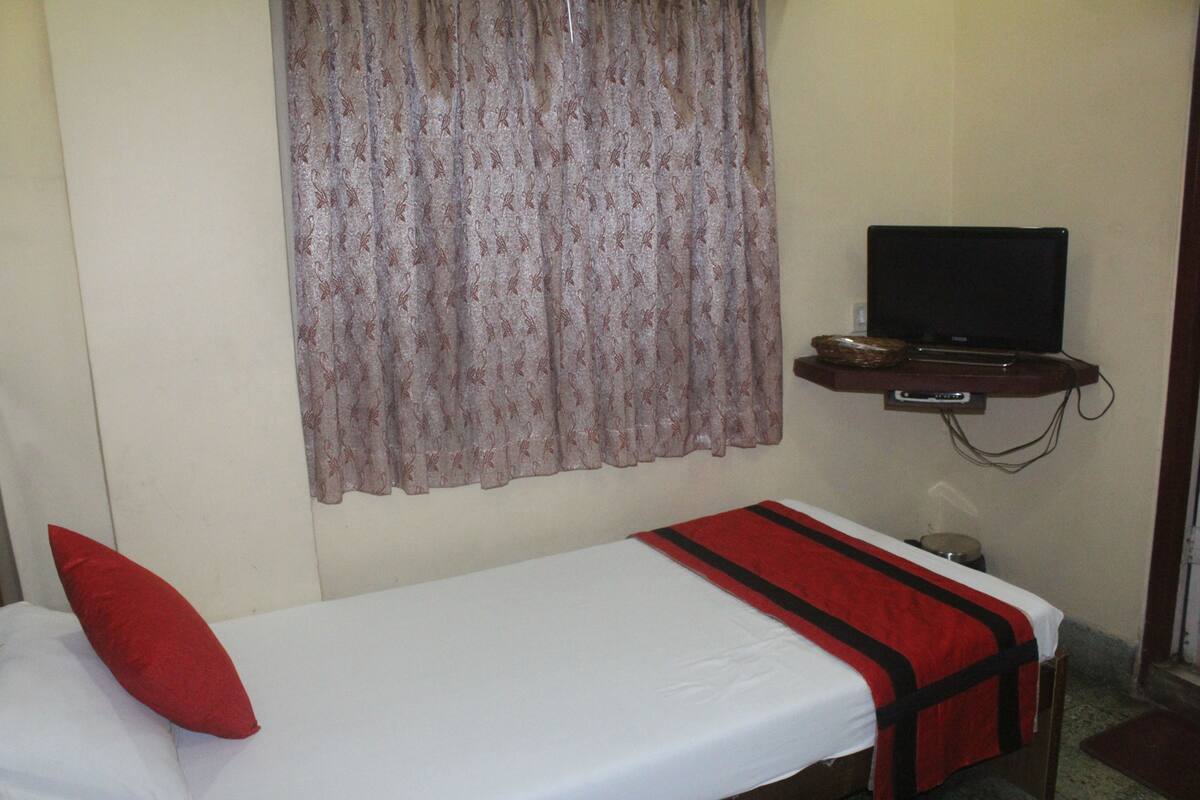 A small, single-bed room with a plain white bedspread, a red pillow, a television on a wooden shelf, and a patterned curtain.