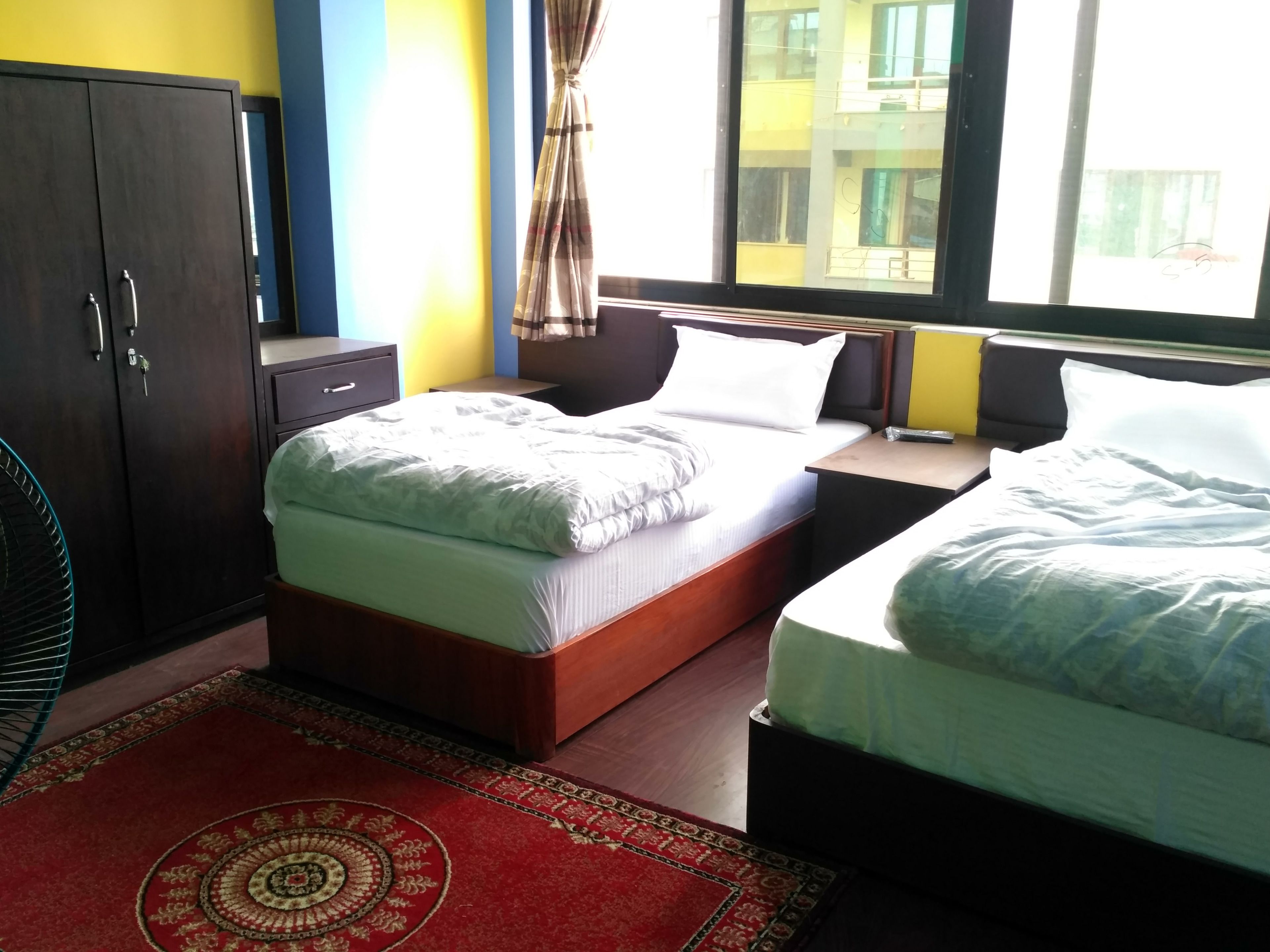 A hotel room with two beds, a wardrobe, a nightstand, and a window with a view of buildings.