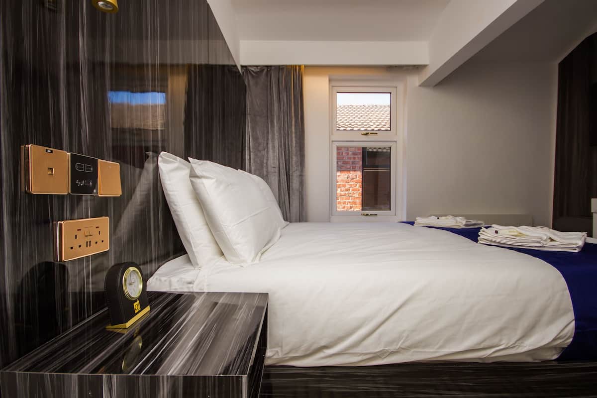 A hotel room with a bed, a bedside table, a clock, and a window with a view of a brick wall.