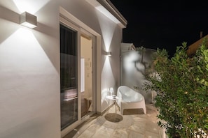 Vue extérieure d’une maison moderne de nuit, avec une entrée bien éclairée, une porte vitrée et un petit espace patio comprenant une chaise et une table blanches.
