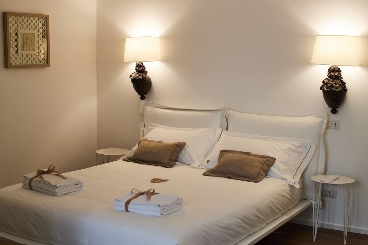 Una cama bien hecha con sábanas blancas y almohadas marrones, dos mesitas de noche con lámparas, y una obra enmarcada en la pared.
