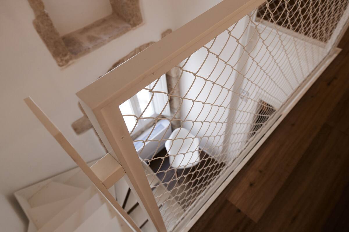 Una habitación con piso de madera, pared blanca y cama blanca con cabecera blanca.