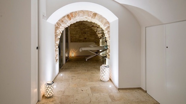A hallway with an arched stone doorway leading to a room with a bed and a small table. There are two wall-mounted lights and a floor lamp. - Iulius Suite & spa (Conversano)