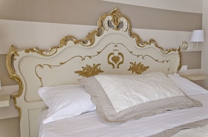 Uma cama com cabeceira elaborada, roupa de cama branca e uma mesa de cabeceira com abajur.
