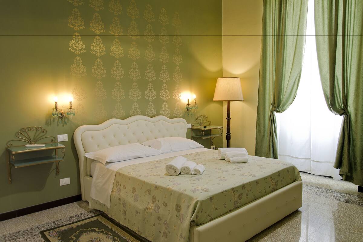 Un lit bien fait, recouvert d’une couvre-lit à motifs floraux, une table de chevet avec une lampe et un mur orné d’un papier peint à motifs.