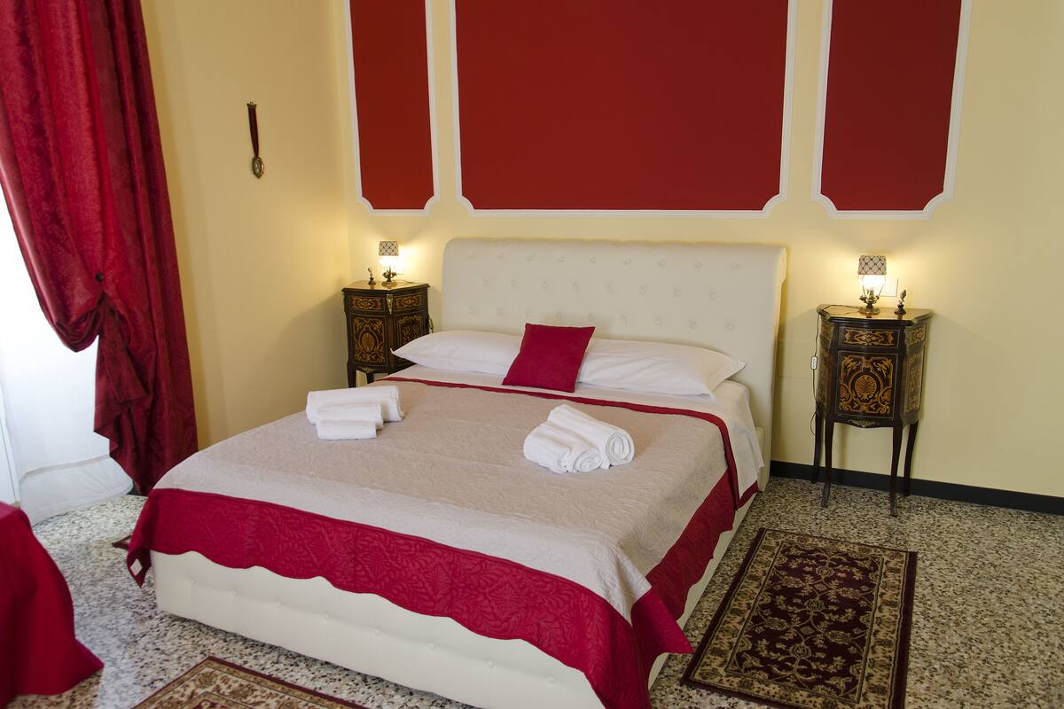 Un lit bien fait, avec du linge de lit blanc et un coussin rouge, flanqué de deux tables de chevet avec des lampes, dans une pièce dotée d’un tapis à motifs et de rideaux rouges.