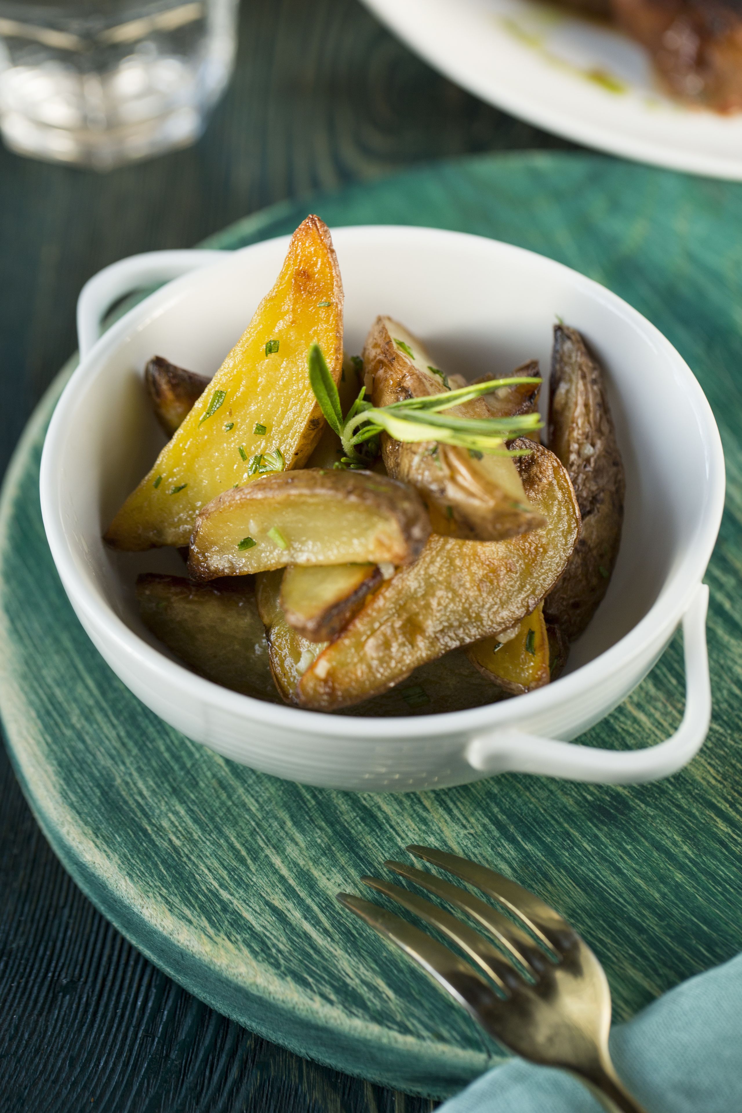 A white bowl of roasted potatoes garnished with herbs, placed on a green textured surface with a fork beside it.