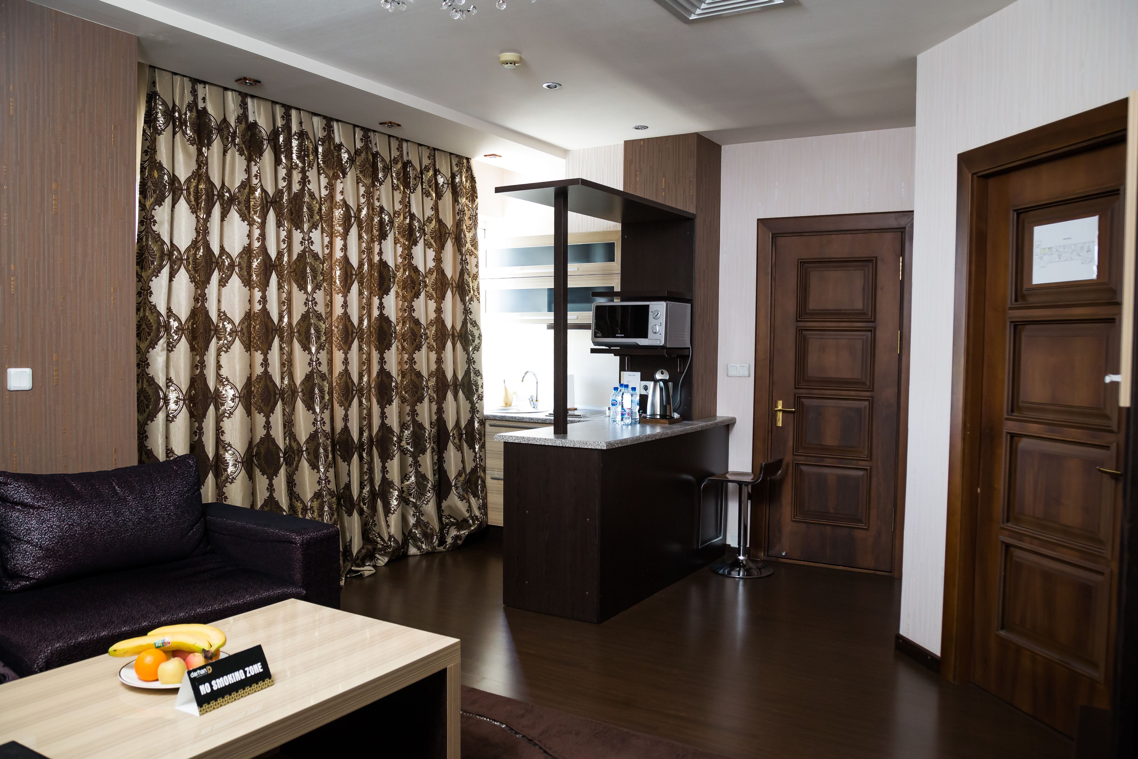 A modern living room with a dark-colored sofa, a small wooden table with fruits, a kitchen area with a microwave, and a wooden door.