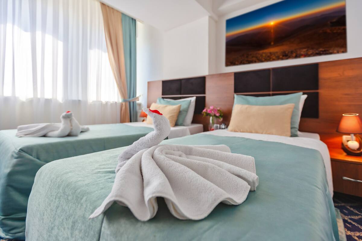 A hotel room with two beds, each with a swan towel arrangement. There is a painting on the wall, a nightstand with a lamp, and a window with curtains.