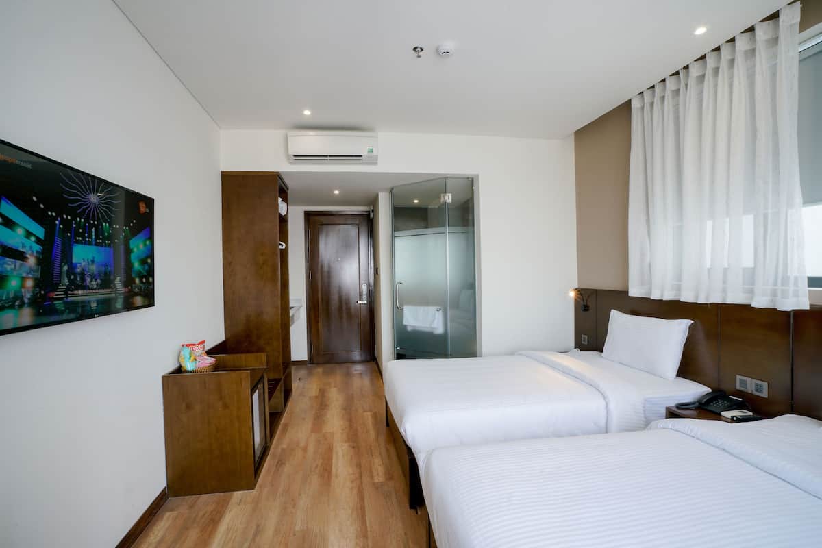 A hotel room with two beds, a wooden desk, a painting, and a glass shower enclosure.