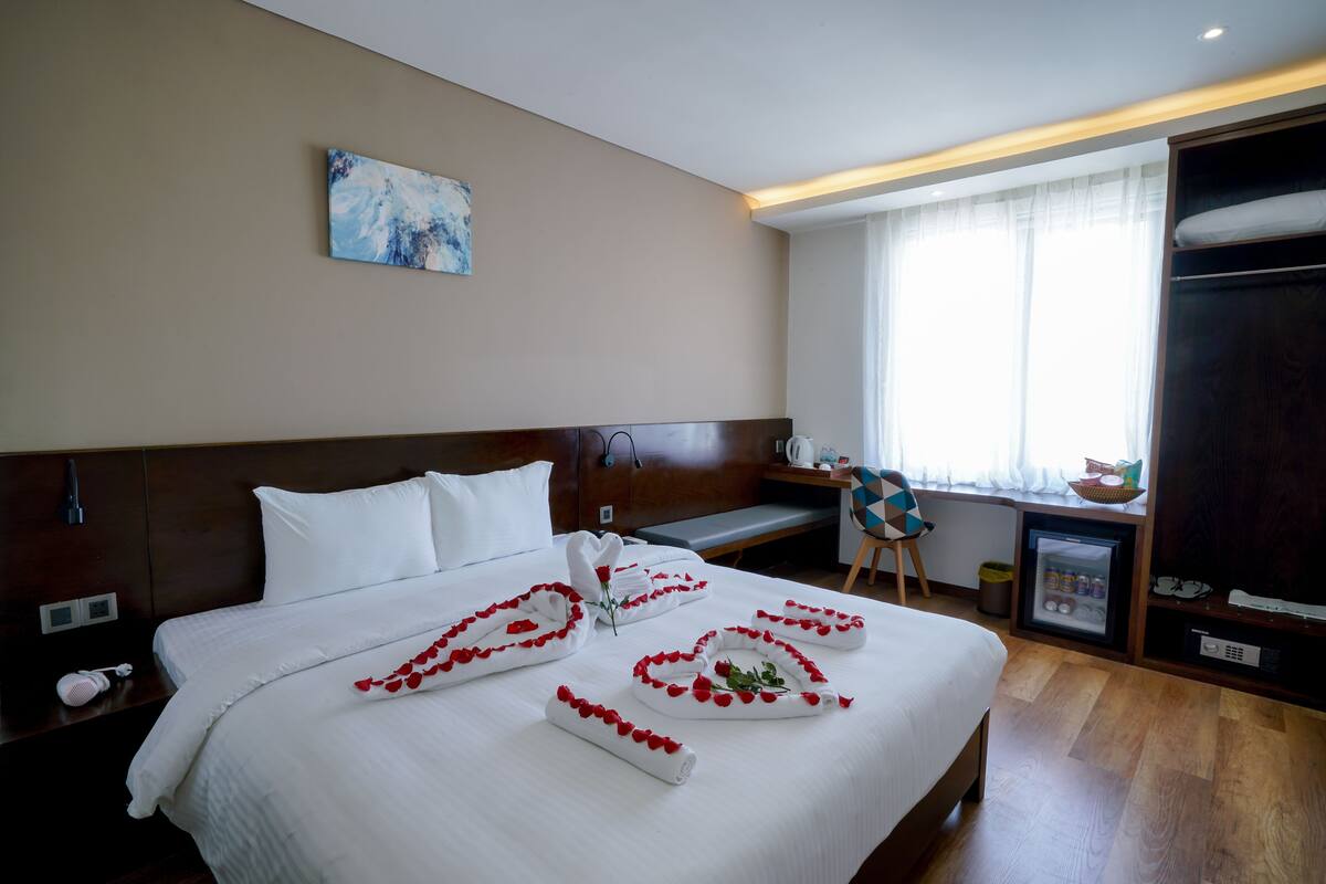 A hotel room with a neatly made bed, a desk with a chair, a small refrigerator, and a window with curtains.