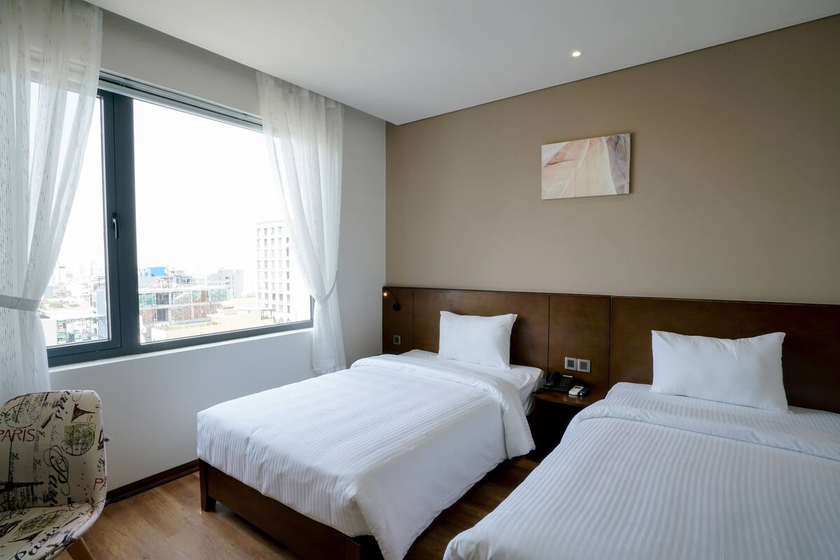 A hotel room with two beds, a window with curtains, a chair, and a painting on the wall.