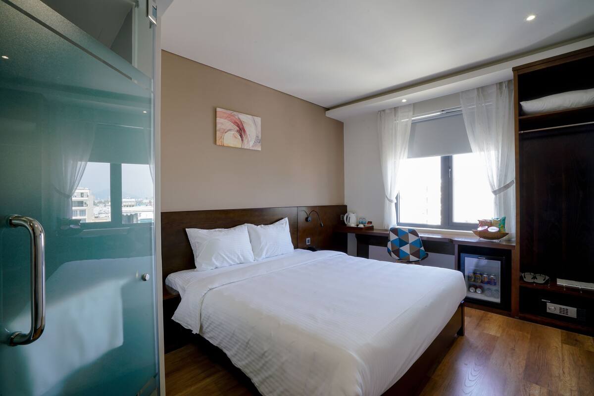 A hotel room with a bed, a bedside table, a small refrigerator, and a window with curtains.