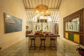 Une cuisine dotée d’un îlot central, de tabourets de bar, de luminaires suspendus en osier, d’un mur en pierre et d’une grande fenêtre agrémentée de plantes.