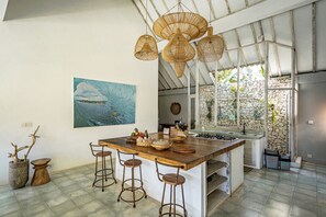 Una cocina con una isla grande, taburetes de bar, lámparas de mimbre colgantes, una pared de piedra y un cuadro de una ola.