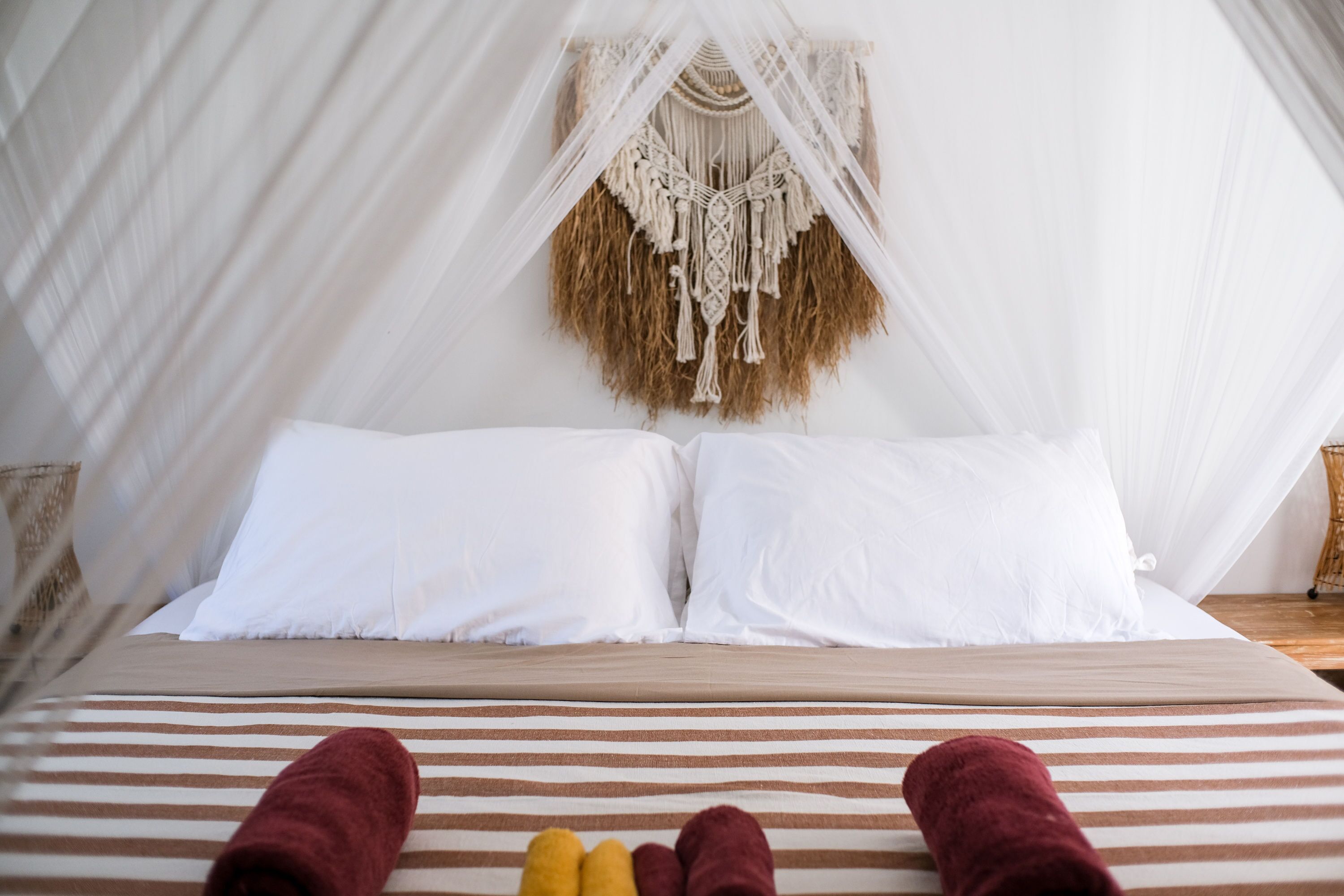 A bed with white pillows and a canopy, a macrame wall hanging, and a striped bedspread.