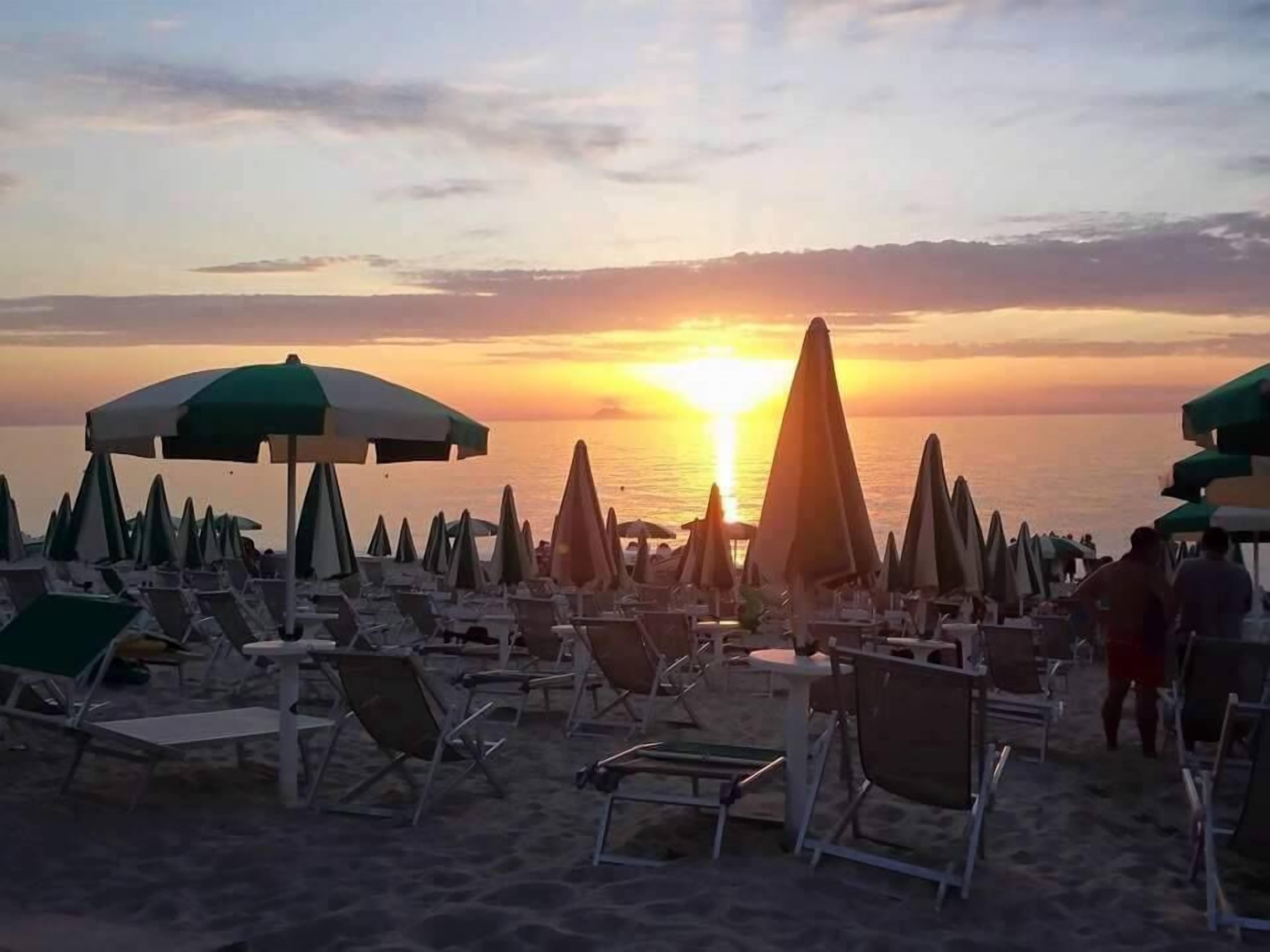 A beach with rows of sun loungers and umbrellas, a sunset in the background, and the ocean visible.