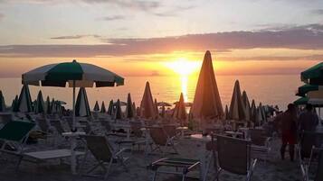 A beach with rows of sun loungers and umbrellas, a sunset in the background, and the ocean visible.