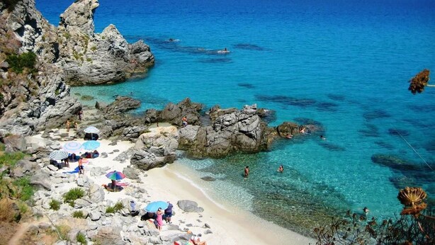 A rocky beach with clear blue water, people swimming, and umbrellas on the shore. - Residenza Mediterranea (Zambrone)