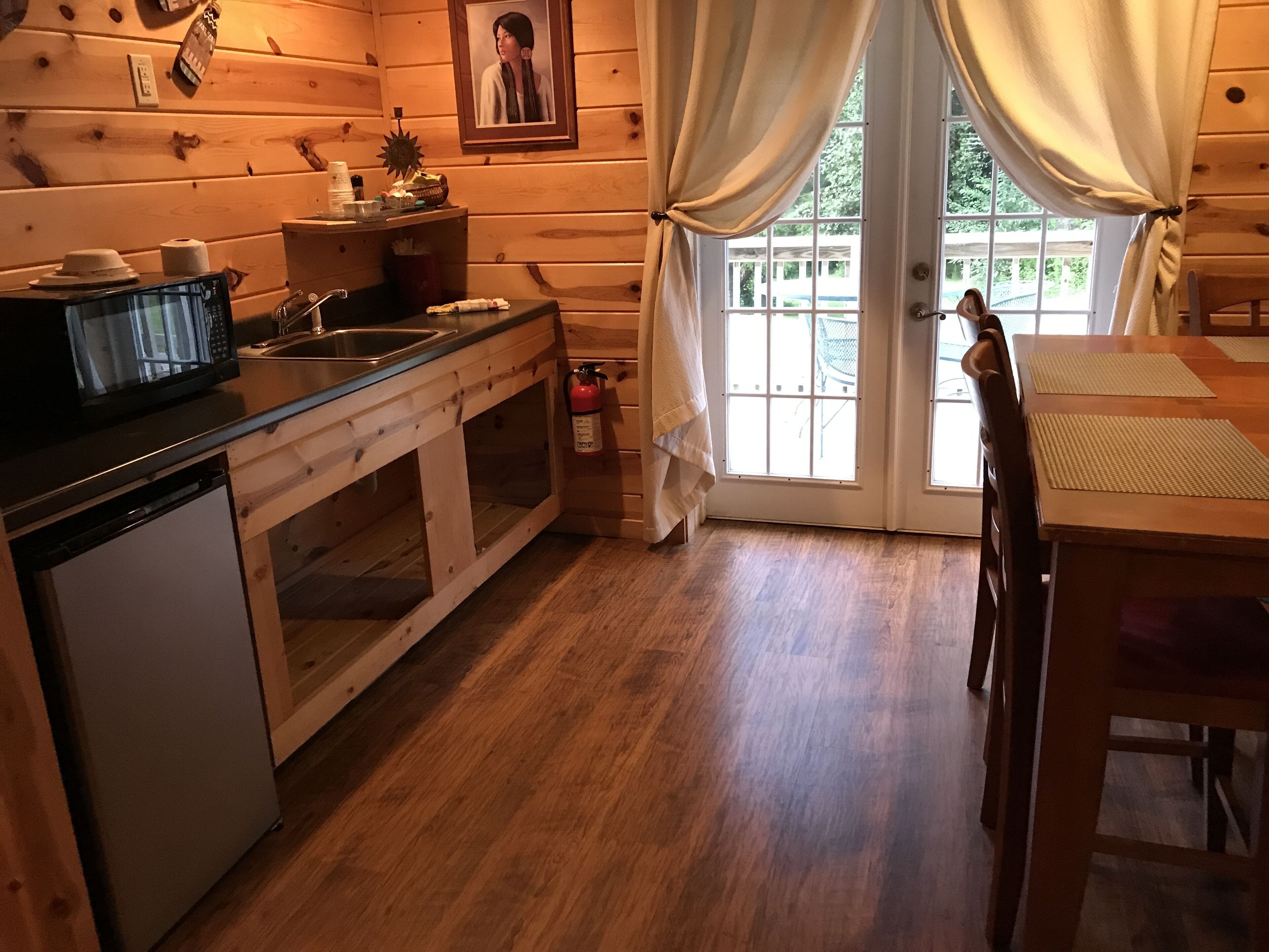 A wooden cabin interior with a kitchen area, a dining table, and a large window with curtains.