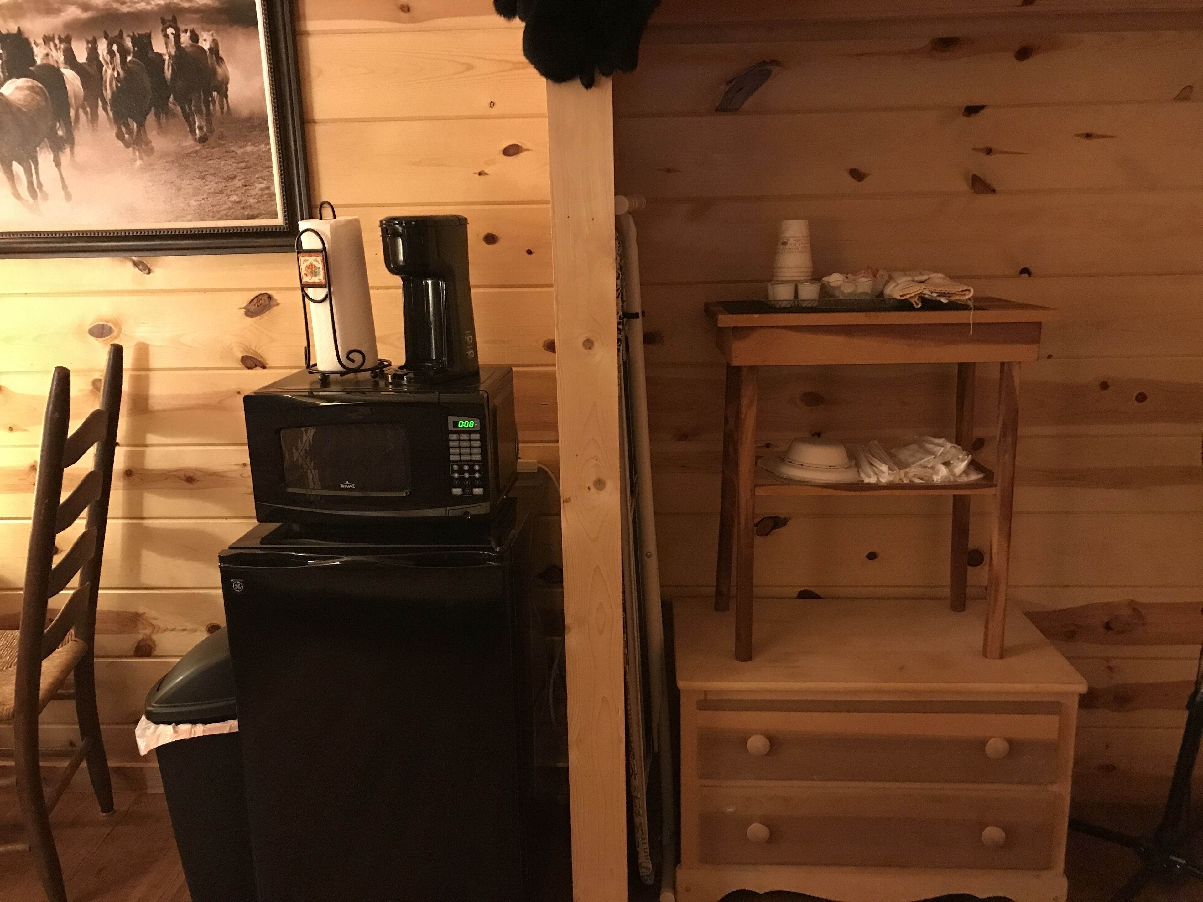 Ett litet, praktiskt köksutrymme med en mikrovågsugn placerad ovanpå ett kylskåp, en kaffebryggare och en liten hylla i trä med koppar och en hatt.
