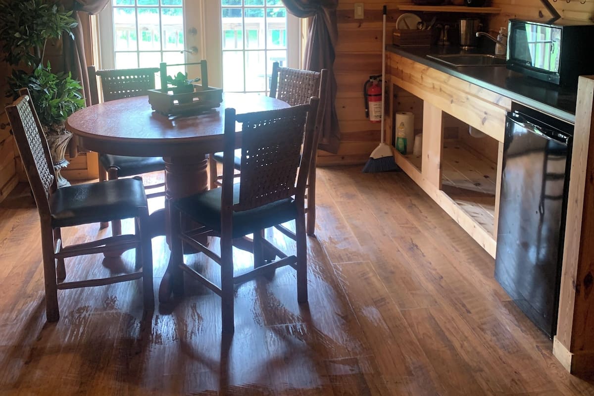 A wooden cabin interior with a dining table, chairs, a kitchen area, and a window with curtains.