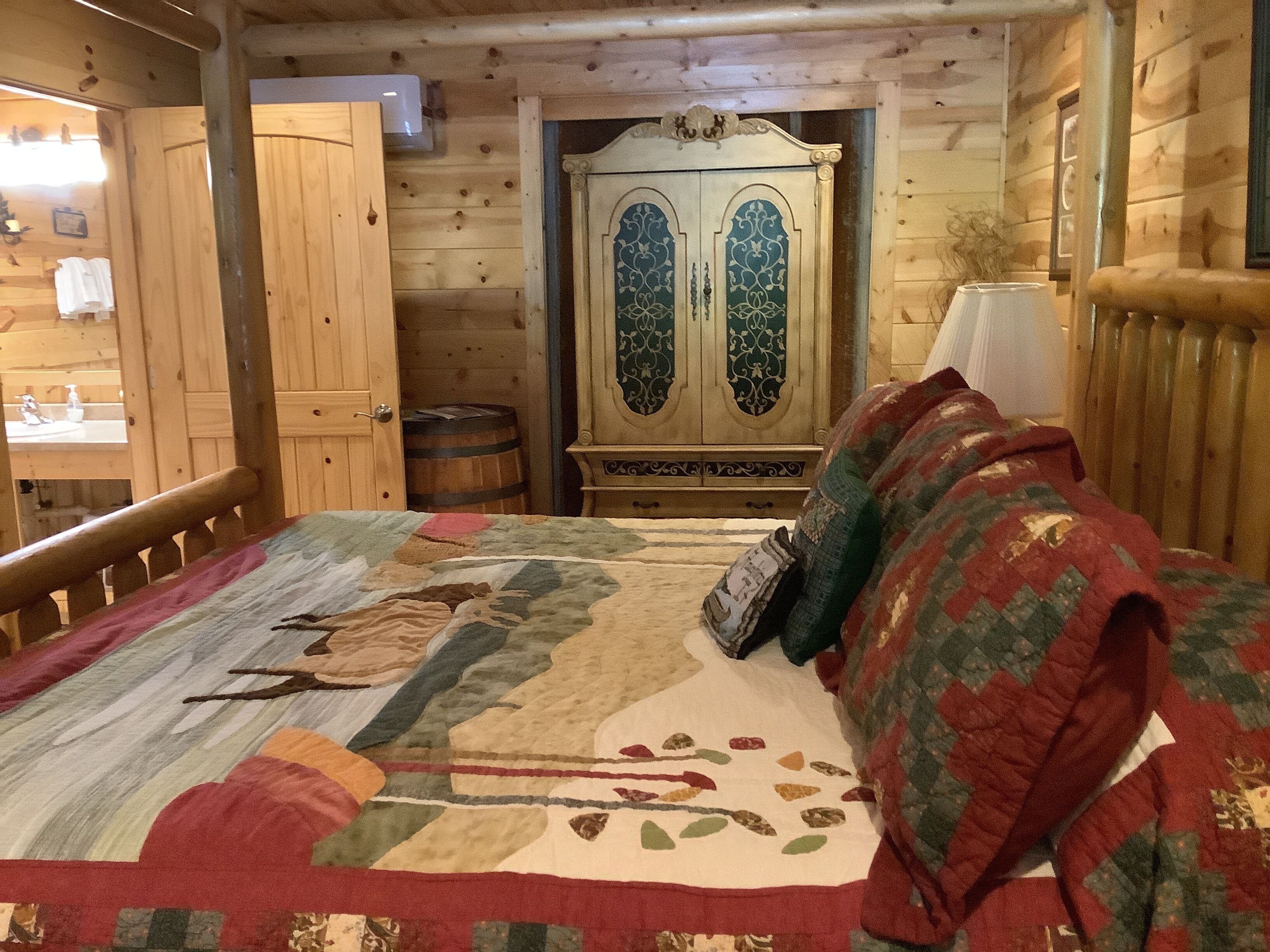 A wooden cabin interior with a bed, a wooden wardrobe, and a small table with a lamp.