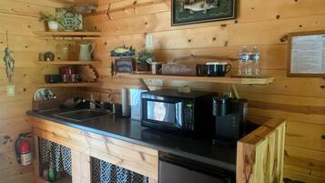 Cocina de cabaña de madera con un lavamanos, microondas, cafetera y refrigerador. Hay estantes con varios artículos y un letrero que dice "Old Bass Lodge Since 1830".