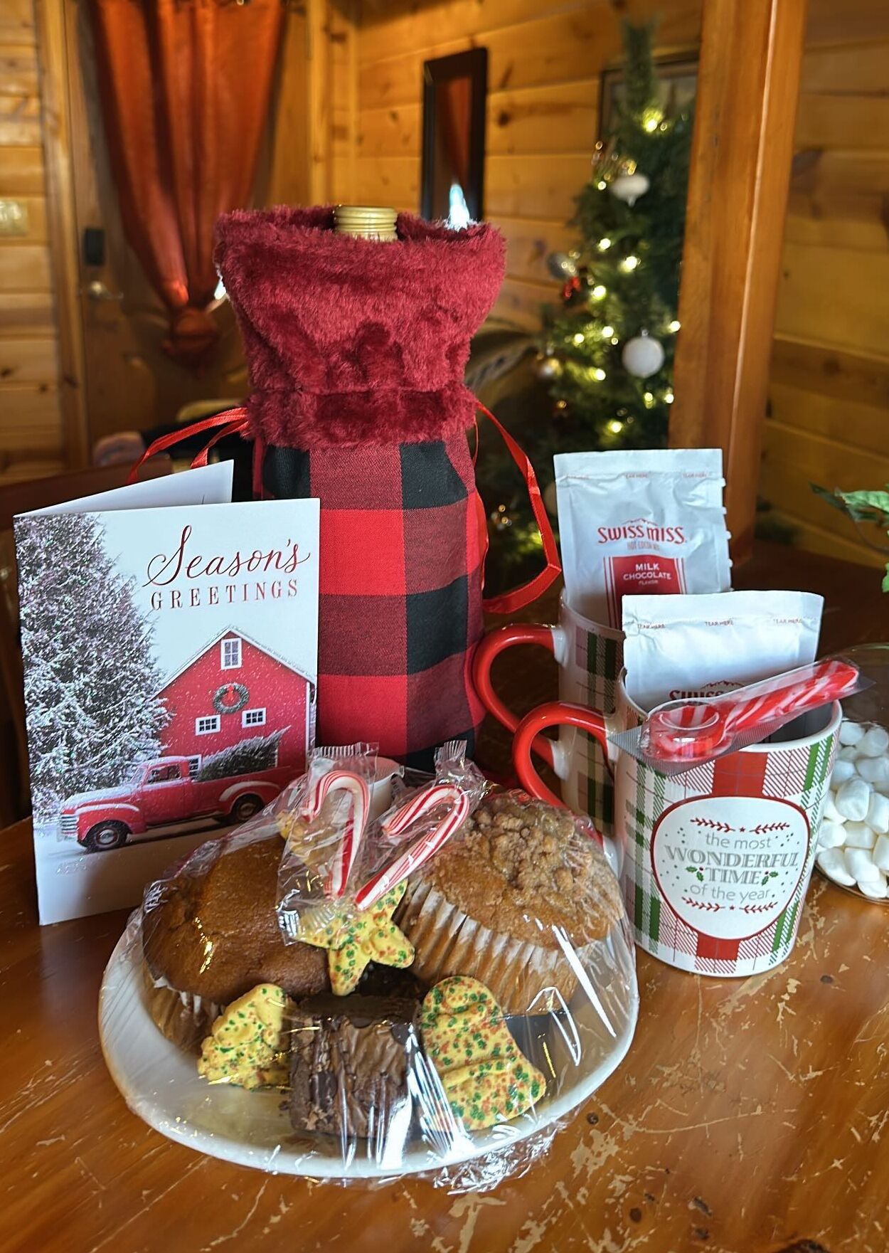 Una mesa festiva con una bolsa a cuadros rojo y negro, una tarjeta con la imagen de un granero rojo, una taza, un plato con galletas y una galleta con forma de estrella, y una bolsa de malvaviscos.