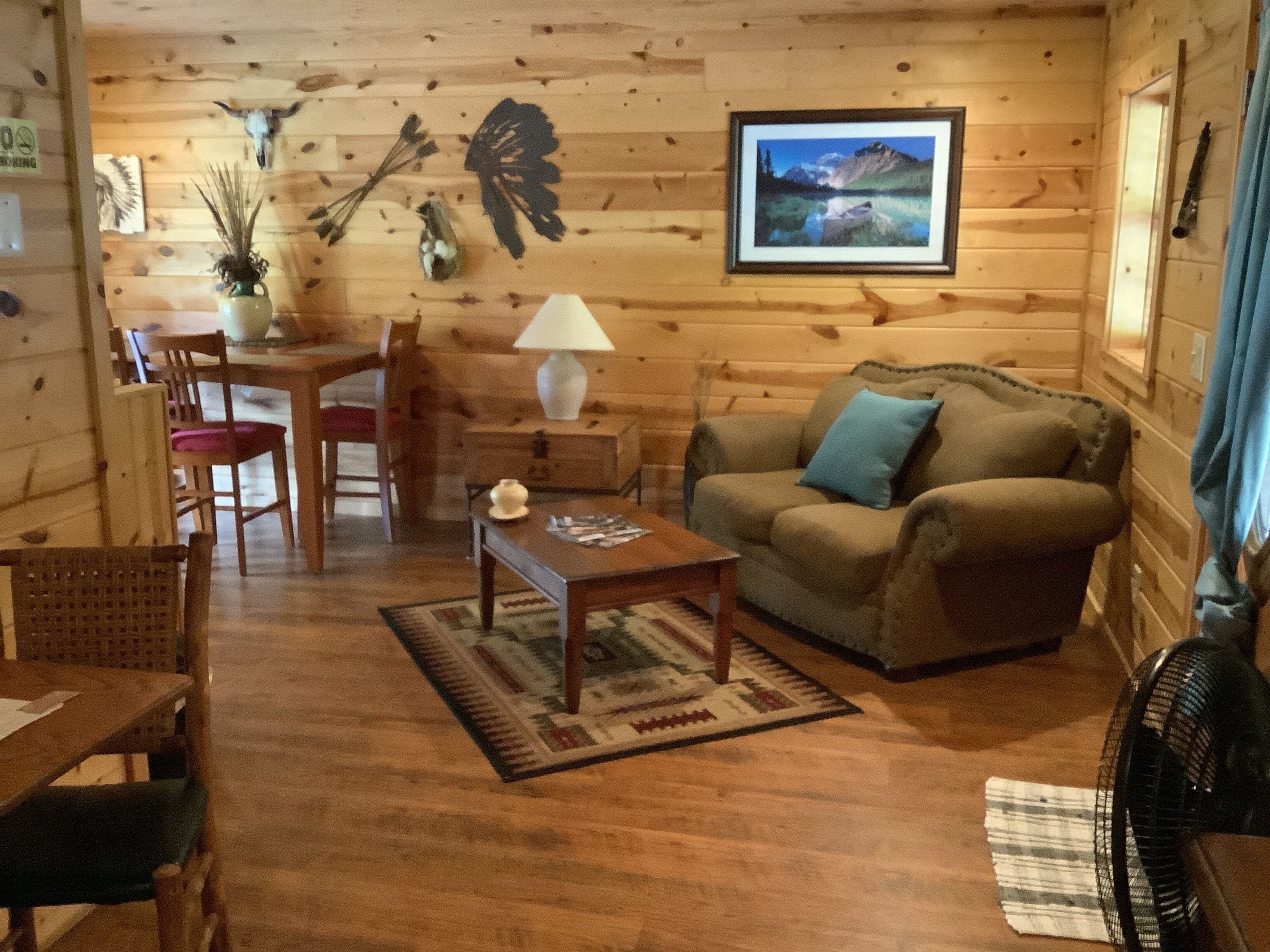 A cozy cabin interior with a wooden floor, a sofa, a dining table with chairs, a lamp, and a framed picture on the wall.