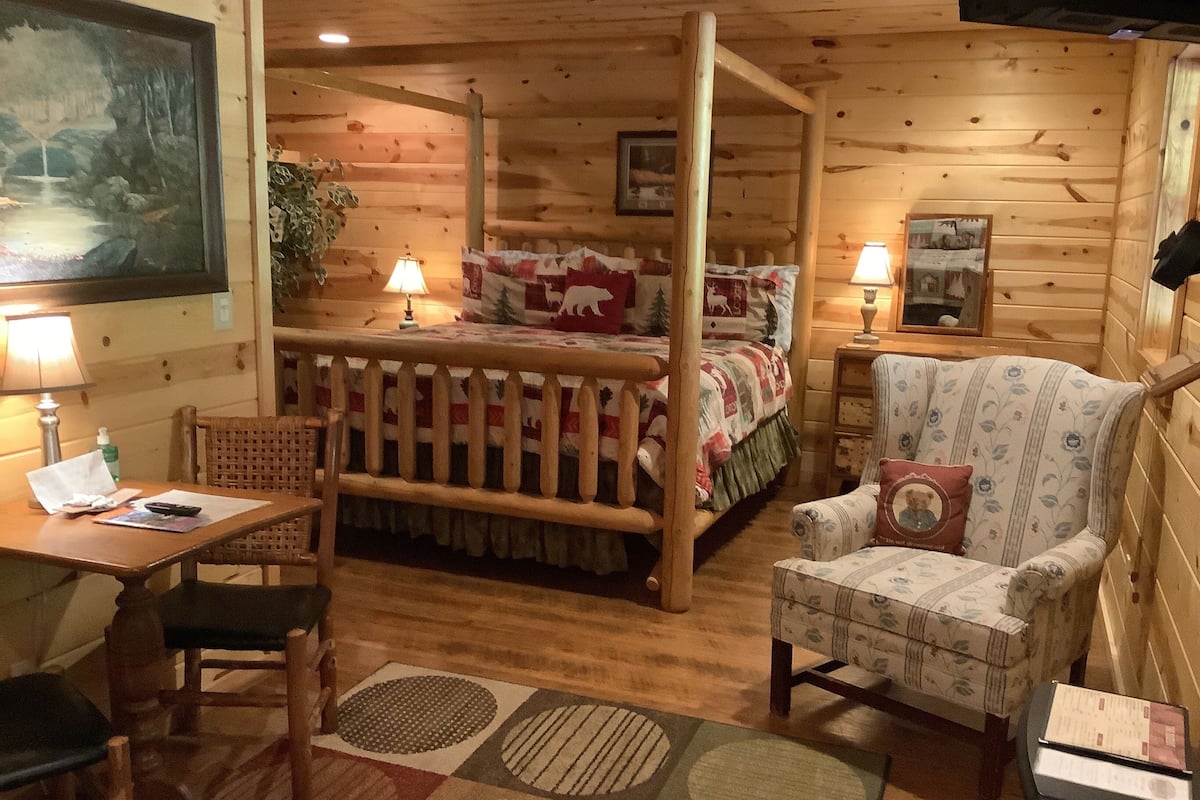 A cozy log cabin room with a four-poster bed, a wooden desk, a chair, and a small table.