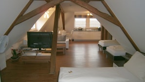 Een slaapkamer op zolder met een televisie, witte meubels, houten balken en een keuken op de achtergrond.