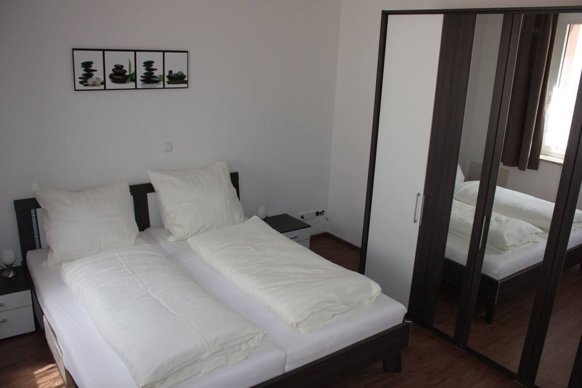 Een tweepersoonsbed met wit beddengoed, een nachtkastje, een bijzettafeltje en een spiegel.