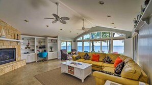 A spacious living room with a stone fireplace, white cabinetry, a yellow sofa, and large windows overlooking a body of water.