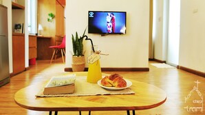 Ein gemütliches Zimmer mit Holztisch, einem Buch, einem Glas Saft und einem Croissant. Ein Fernseher ist an der Wand angebracht und eine Pflanze steht in der Ecke.