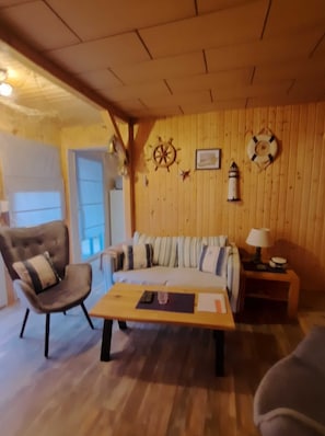 A cozy living room with a wooden ceiling, a sofa with striped cushions, a wooden coffee table, a chair, and a small table with a lamp. - Bungalow 2 - Apartment and bungalow in Trassenheide (Zinnowitz)