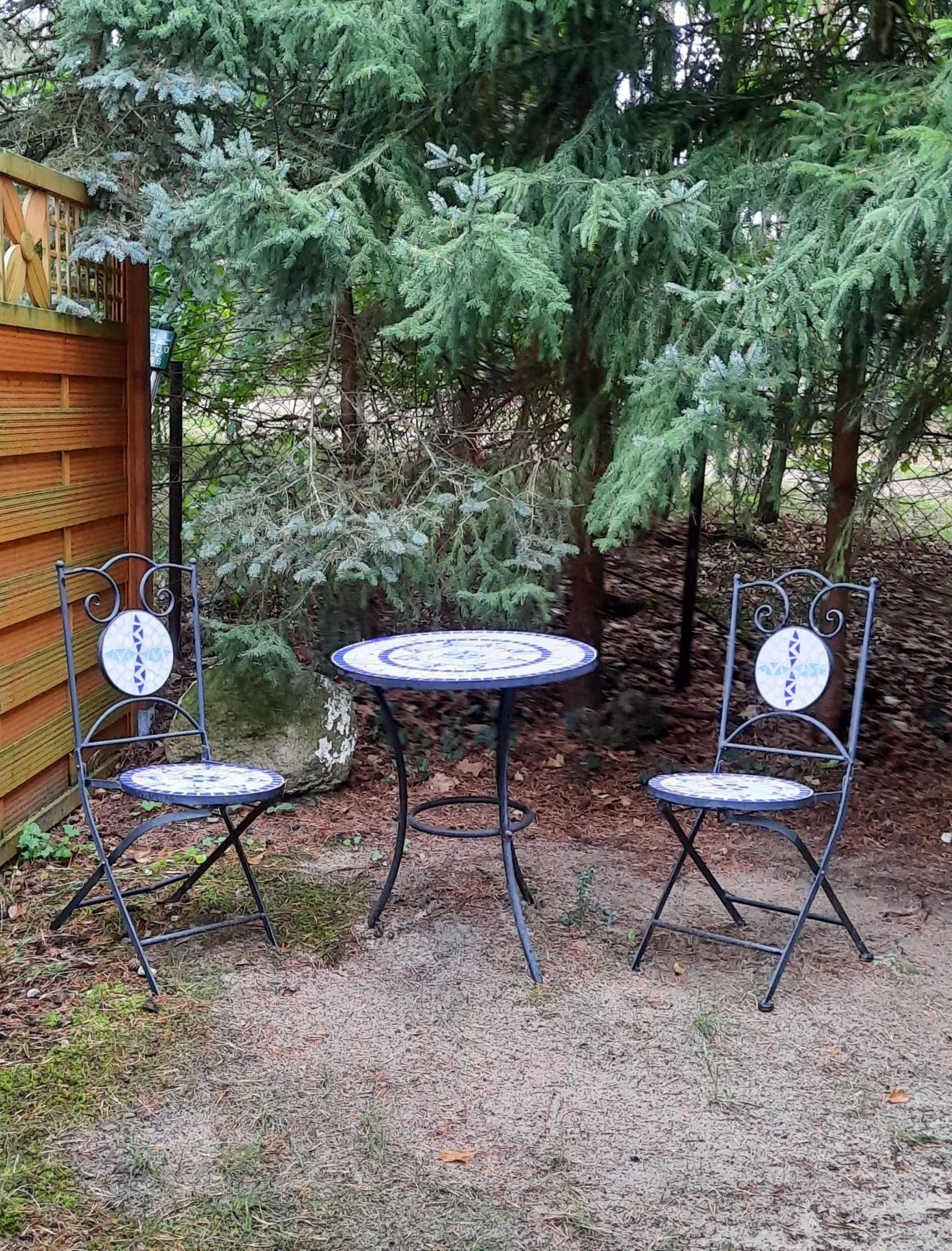 Two metal chairs with circular tables, set on a gravel path in a garden with a wooden fence and evergreen trees in the background.