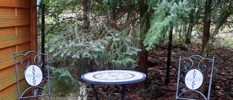 Two metal chairs with circular tables, set on a gravel path in a garden with a wooden fence and evergreen trees in the background.
