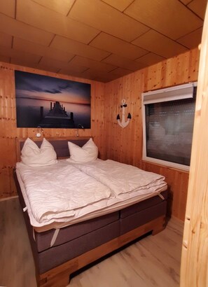 A wooden-paneled room with a bed, a painting of a sunset over a pyramid, and a window with a view of the city. - Bungalow 1 - Apartment and bungalow in Trassenheide (Zinnowitz)