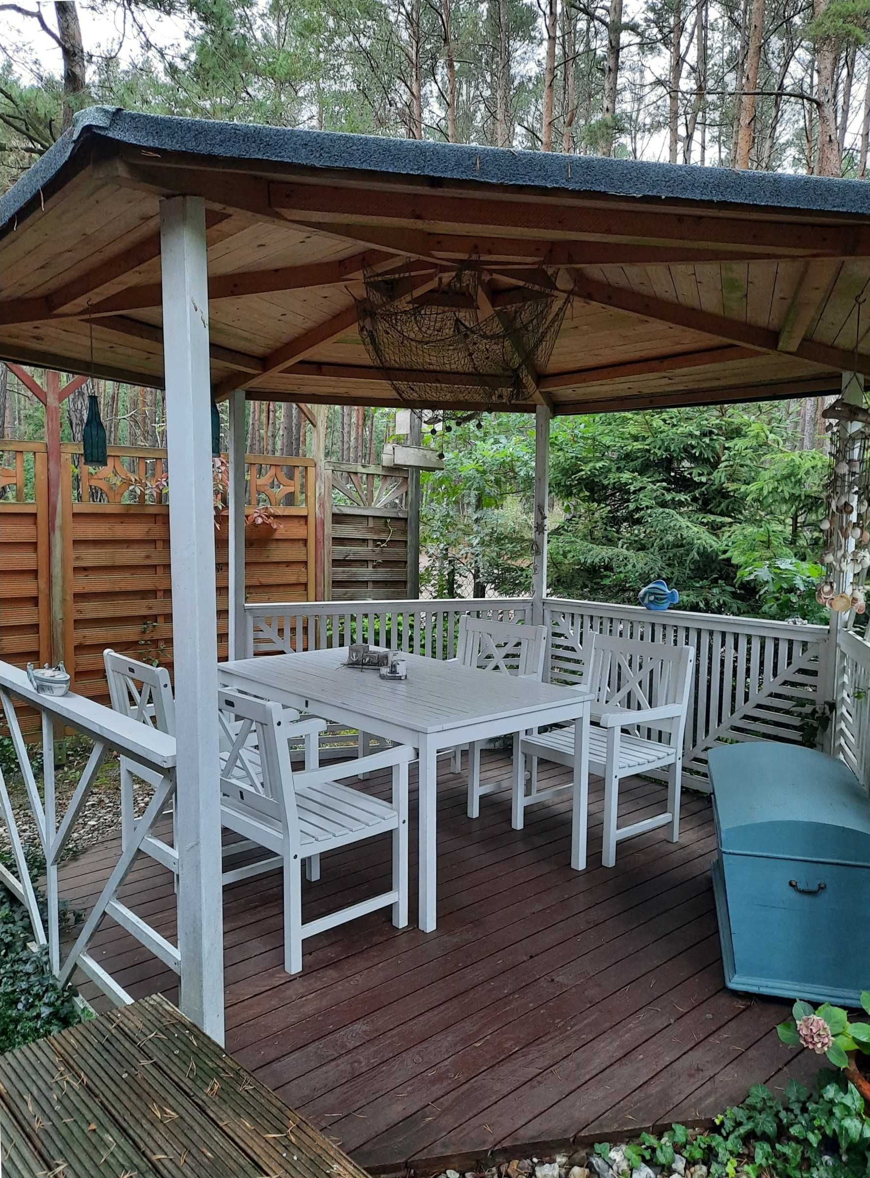 A covered outdoor patio with a wooden table and white chairs, surrounded by a wooden deck and a white fence.