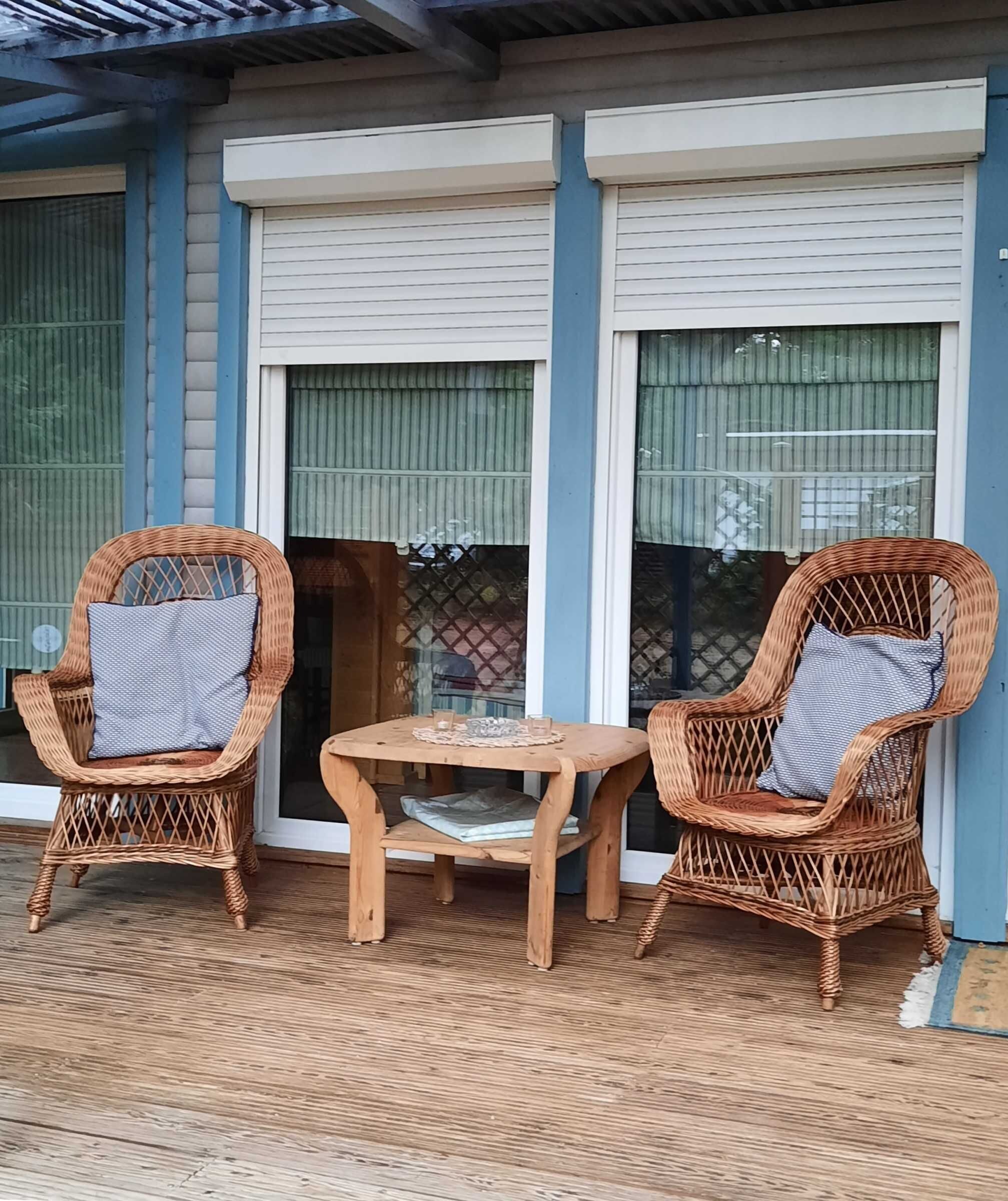 A covered outdoor seating area with two wicker chairs and a small wooden table, featuring a view of a building with large windows and a blue exterior.