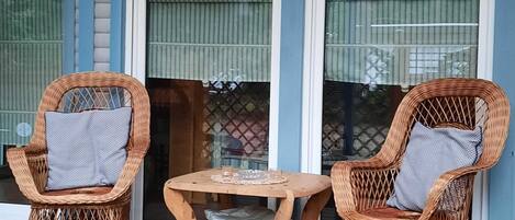 A covered outdoor seating area with two wicker chairs and a small wooden table, featuring a view of a building with large windows and a blue exterior.