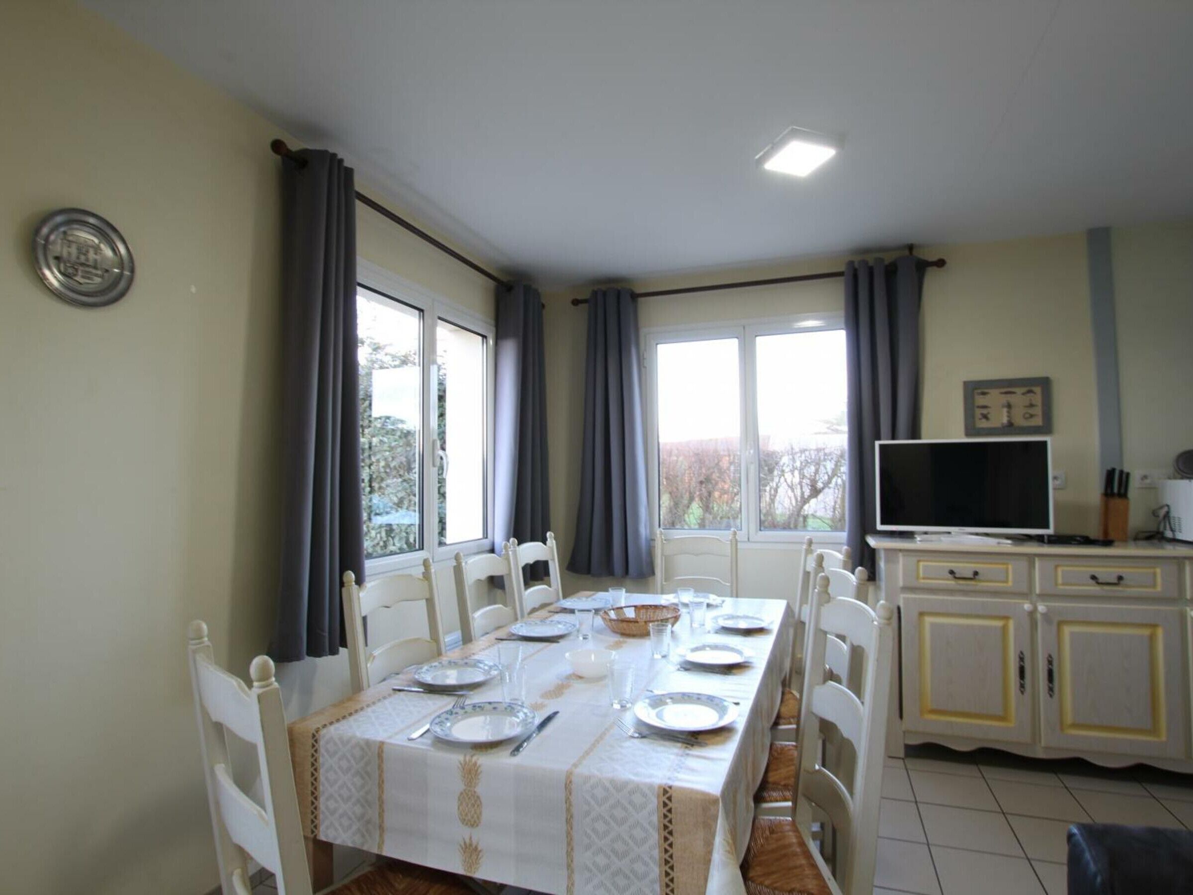 A dining area with a long table set for four, white chairs, a white cabinet with a television, and a window with curtains.