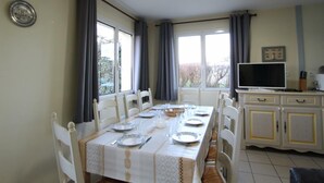 Ein Essbereich mit einem langen Tisch, gedeckt für vier Personen, weißen Stühlen, einem weißen Schrank mit Fernseher und einem Fenster mit Vorhängen.