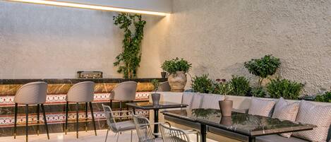 A modern outdoor seating area with a glass table and metal chairs, surrounded by a textured wall and potted plants.