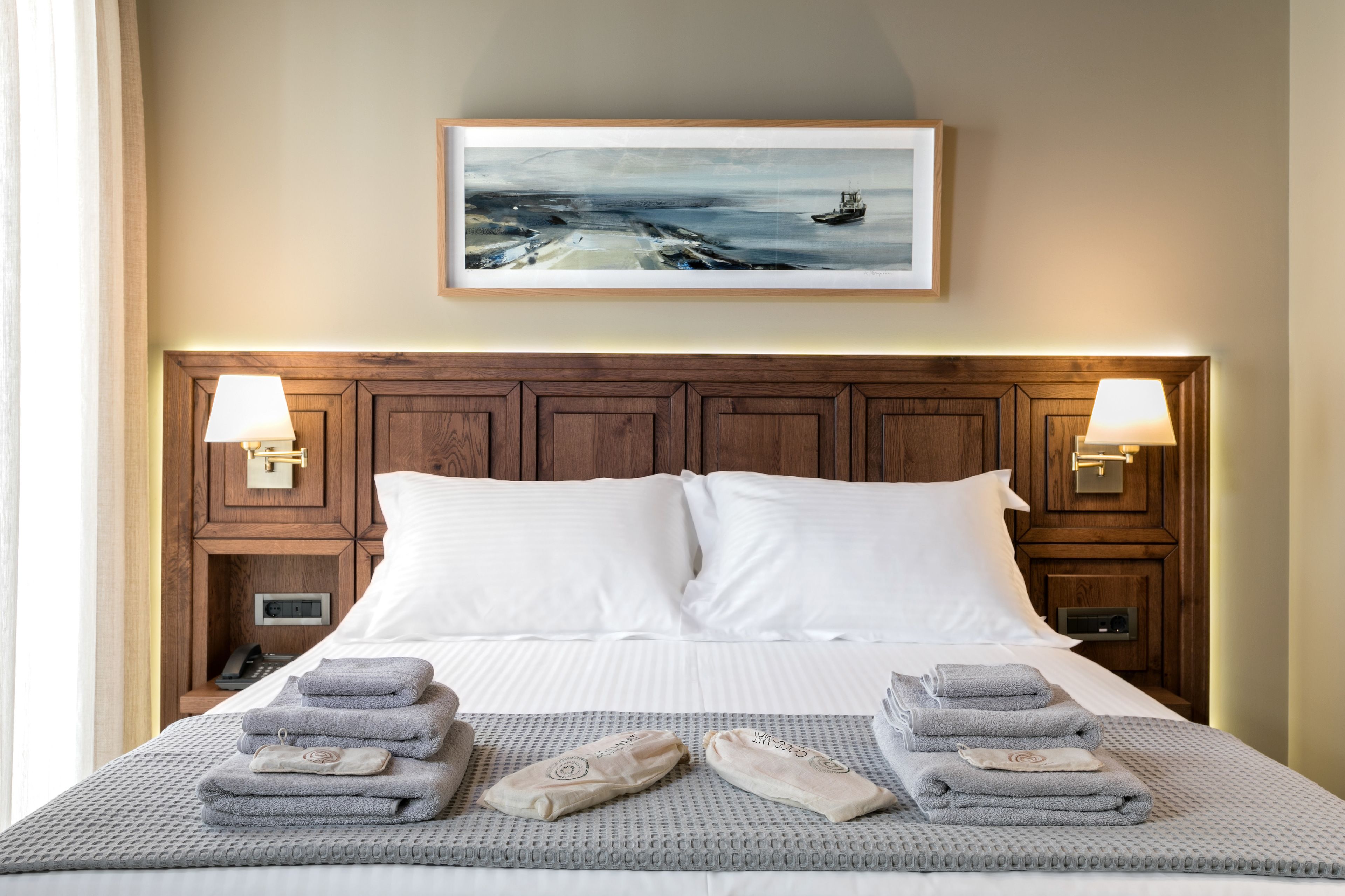 A neatly made bed with white linens, a grey textured blanket, and folded towels. There is a framed painting above the bed depicting a seascape.