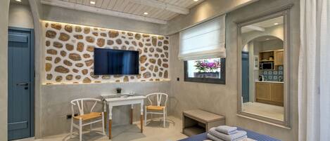 Un dormitorio con pared de piedra, televisión, escritorio, dos sillas, cama con toallas y baño visible a través de un arco.