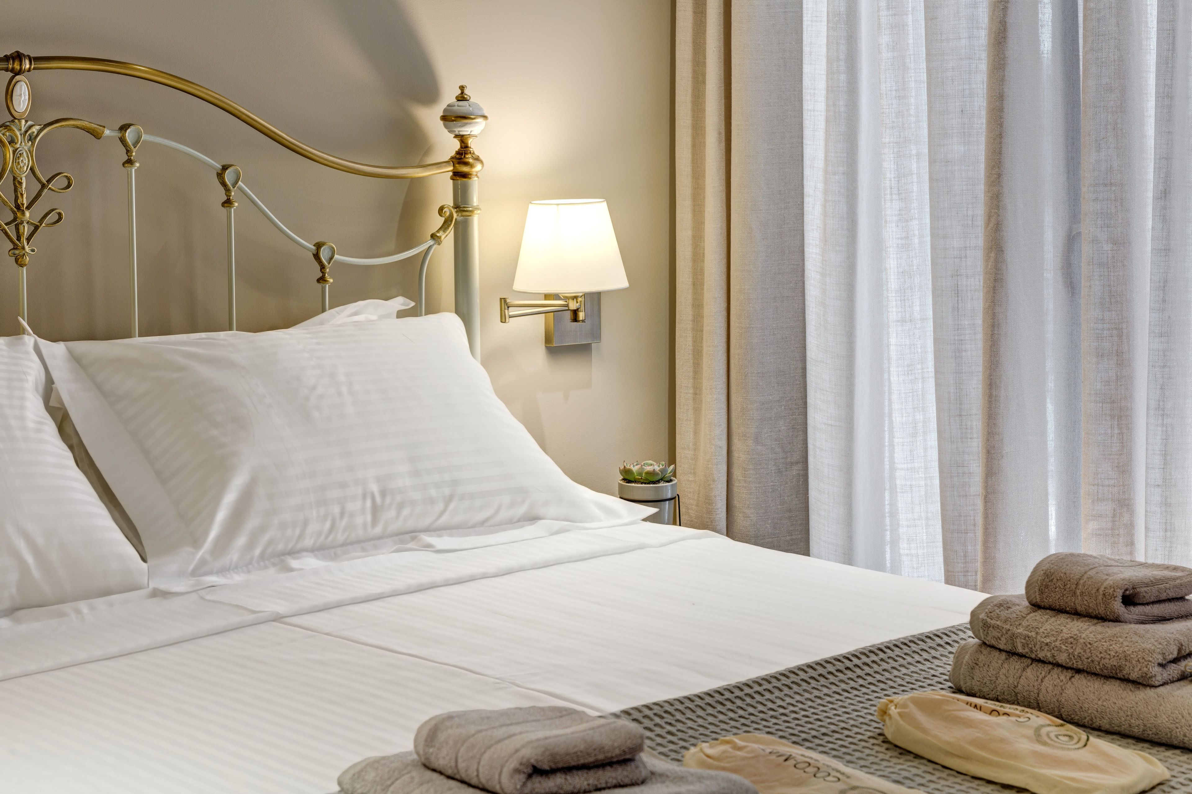 A neatly made bed with white linens, a gold ornate headboard, a wall-mounted lamp, and a small potted plant beside the bed.