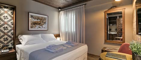 A neatly made bed with white linens and folded towels, a wooden ceiling, a framed picture on the wall, a small table with a plant, and a mirror reflecting a kitchen area.